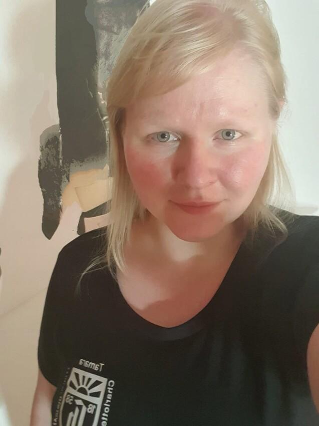 Porträt einer Person mit blonden Haaren, die ein schwarzes T-Shirt trägt, vor einer künstlerischen Wandgestaltung.