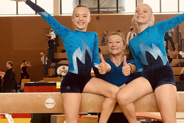 Zwei Turnerinnen in blauen Trikots posieren freudig auf einem Schwebebalken mit Trainerin, alle geben Daumen hoch.