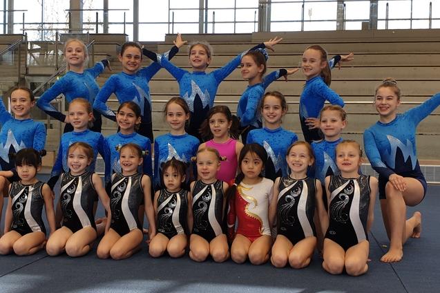 Gruppe von 20 Turnerinnen in blauen und schwarzen Gymnastikanzügen auf einer Mattenfläche, lächelnd und posierend.