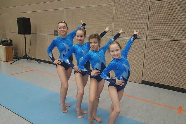 Vier Mädchen in blauen Gymnastikanzügen posieren lächelnd auf einer Gymnastikmatte in einer Sporthalle.