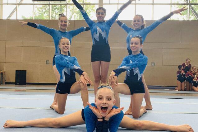 Sechs Turnerinnen in blauen Anzügen posieren im Wettkampf auf einer Turnmatte, eine liegt im Spagat vorne.