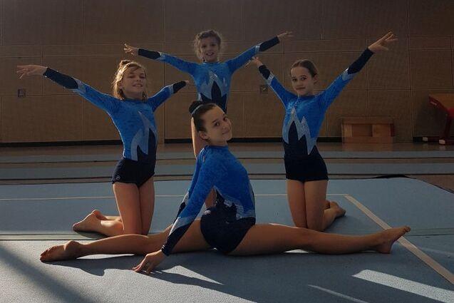 Vier Turnerinnen in blauen Sportanzügen posieren auf einer Turnerfläche, eine sitzt im Spagat, die anderen stehen.