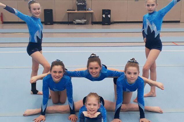 Sechs junge Turnerinnen in blauen Trikots posieren kreativ auf einem Gymnastikboden.