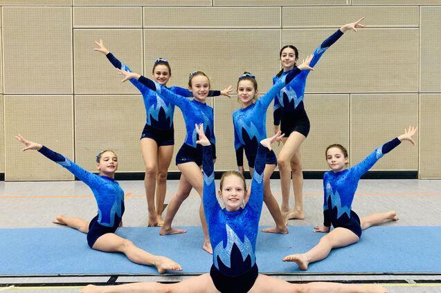 Sechs Turnerinnen in blauen Trikots zeigen eine Pose auf einer Gymnastikmatte in einer Halle.