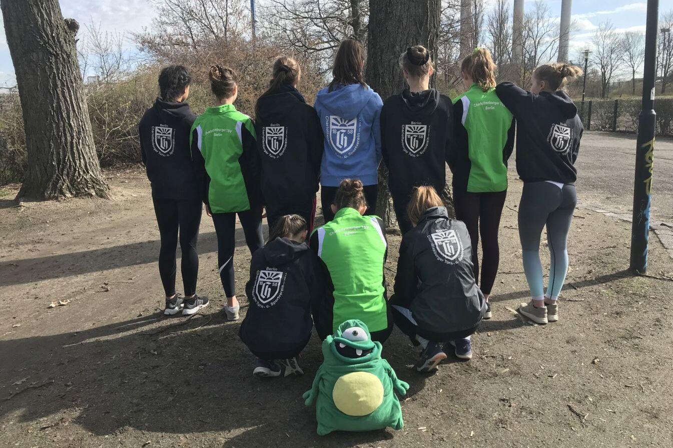 Gruppe von Sportlerinnen, die von hinten stehen, in Jacken mit Logos, eine sitzt in einem grünen Outfit.