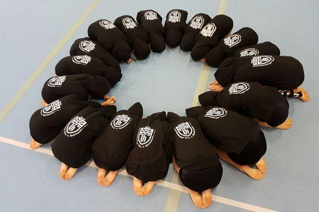 Eine Gruppe von Personen in schwarzen Kapuzenpullis sitzt hockend in einem Kreis auf einem blauen Boden.