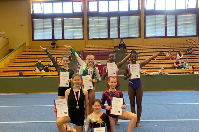 Gruppe junger Turnerinnen präsentiert stolz Urkunden und Medaillen auf einer Gymnastikmatte in einer Sporthalle.