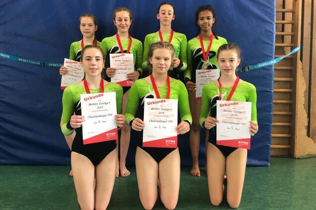 Sechs junge Turnerinnen mit Medaillen halten Urkunden des Berliner Turnliga Wettkampfs 2019 in einer Sporthalle.