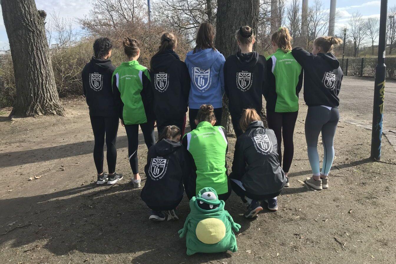 Gruppe von Sportlerinnen, die von hinten stehen, in Jacken mit Logos, eine sitzt in einem grünen Outfit.