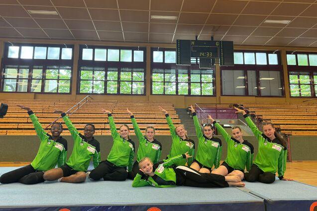 Gruppe junger Turnerinnen in grünen Trainingsjacken posiert auf einem Turnboden in einer Sporthalle.