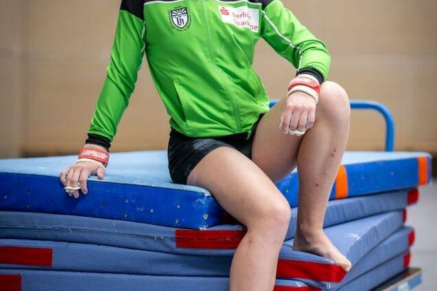 Junge Sportlerin in grüner Trainingsjacke sitzt lächelnd auf einem blauen Schaumstoffblock in einem Sportbereich.