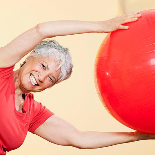 Ältere Frau in Sportkleidung streckt sich lächelnd zu einem großen roten Gymnastikball.