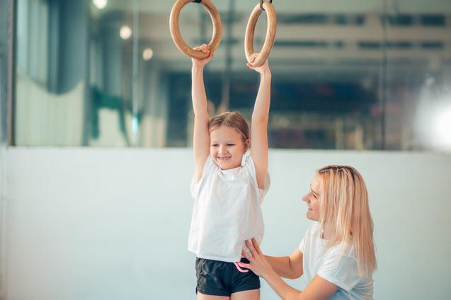 Ein Mädchen hängt an Holzringen, während eine Trainerin sie unterstützt und anlächelt in einer Sporthalle.