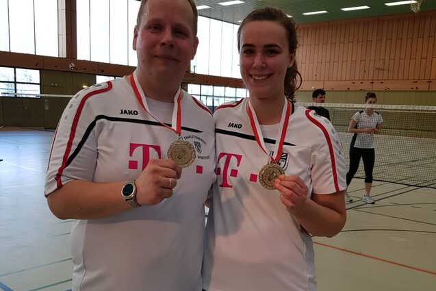 Zwei Sportler in weißen Trikots halten Medaillen in einer Sporthalle, Hintergrund zeigt Badmintonfeld und weitere Personen.