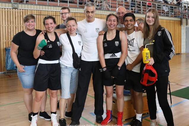 Gruppe von fünf Boxsportlern und Trainern lächelt in einer Sporthalle, im Hintergrund Zuschauer auf Tribüne.