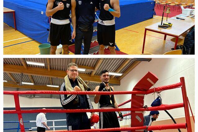 Drei Boxer posieren mit Trainer in einer Sporthalle neben einem Boxring und Tisch, während Zuschauer im Hintergrund sitzen.