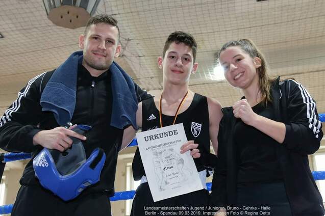 Drei Personen posieren in einem Boxring, eine hält eine Urkunde für den ersten Platz in einem Wettkampf.