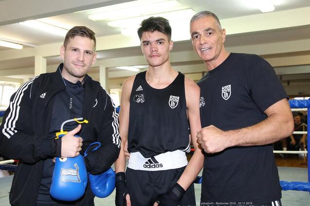 Drei Boxer stehen in einem Trainingsraum: Zwei Trainer und ein Kämpfer in schwarzer Sportkleidung mit Boxhandschuhen.