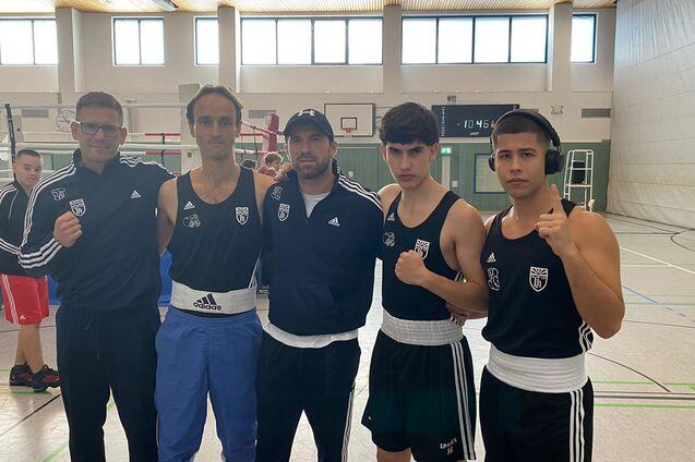 Fünf Boxer in Trainingsanzügen posieren in einer Turnhalle, der Boxring ist im Hintergrund sichtbar.