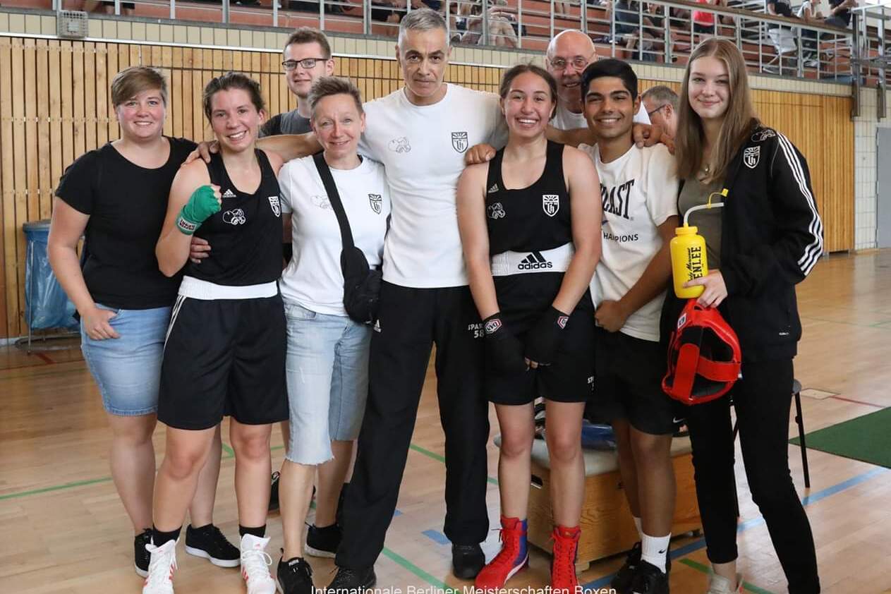 Gruppe von fünf Boxsportlern und Trainern lächelt in einer Sporthalle, im Hintergrund Zuschauer auf Tribüne.