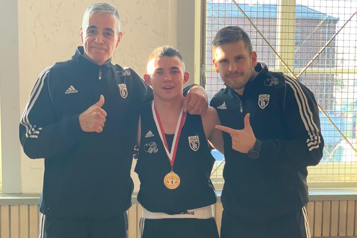 Drei Sportler stehen in einem Raum, der Medaillenhalter trägt eine Goldmedaille und lächelt stolz.