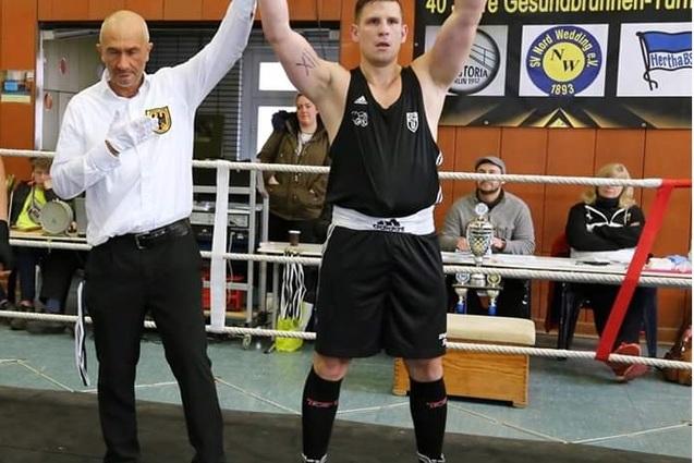 Boxer in schwarzem Trikot hebt die Arme jubelnd, während ein Schiedsrichter den Sieg verkündet. Publikum im Hintergrund.