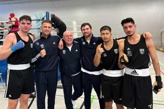 Fünf Boxer in schwarzen Trikots posieren gemeinsam in einer Sporthalle nach einem Wettkampf.