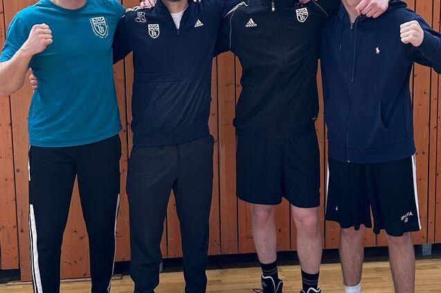 Vier Personen posieren lächelnd in Sportbekleidung vor einer Holzwand in einer Sporthalle.