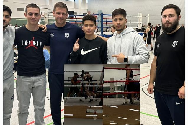 Fünf Personen posieren lächelnd vor einer Boxsport-Veranstaltung, im Hintergrund sind Boxringe zu sehen.