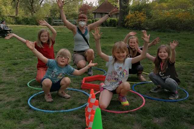 Sechs Kinder und eine Erwachsene posieren fröhlich im Park, umringt von Hula-Hoop-Reifen und bunten Pylonen.