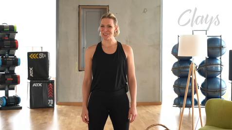 Frau in schwarzem Sportoutfit steht in einem Fitnessraum mit Übungsgeräten und einer Lampe im Hintergrund.