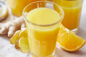 Glas mit Ingwer-Orangen-Shot, umgeben von Ingwerstücken und Orangenscheiben auf einem Holzbrett.