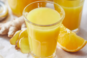 Glas mit Ingwer-Orangen-Shot, umgeben von Ingwerst&uuml;cken und Orangenscheiben auf einem Holzbrett.