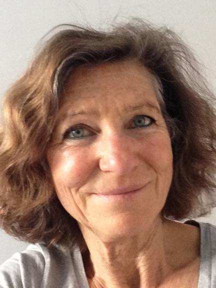 Portrait einer lächelnden Frau mit kurzem, lockigem Haar vor neutralem Hintergrund. Sie trägt ein graues Oberteil.