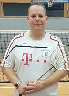 Mann mit Badmintonschläger steht in einem Sporthallenbereich vor einem Badmintonnetz.
