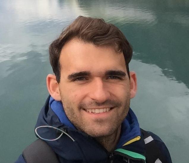 Lächelnder Mann mit kurzen Haaren, stehend vor einem Gewässer, trägt eine Jacke und hält sein Handy in der Hand.