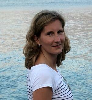 Frau mit lockigem Haar lächelt leicht vor einem blauen Wasserhorizont, trägt ein gestreiftes T-Shirt.