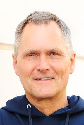 Porträt eines Mannes mit kurzen grauen Haaren, der in einem blauen Kapuzenpullover lächelt. Heller Hintergrund.