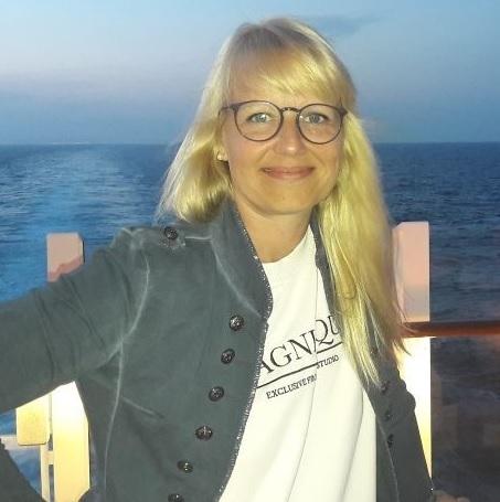 Frau mit Schulterlangen blonden Haaren und Brille steht vor einem Meer im Abendlicht. Sie trägt einen grauen Blazer.