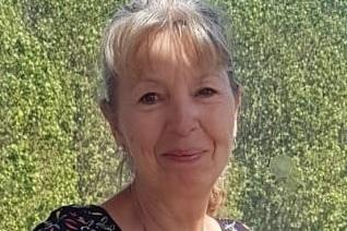 L&auml;chelnde Frau mit blonder, halblanger Haarfrisur, tr&auml;gt ein gemustertes Oberteil vor gr&uuml;ner Kulisse.