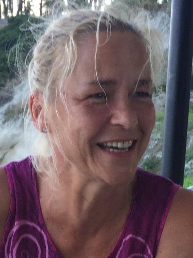 Lächelnde Frau mit blonden, lockigen Haaren, die ein lila Oberteil trägt, vor einer natürlichen Kulisse.