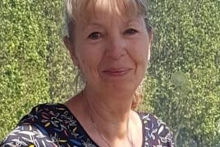 L&auml;chelnde Frau mit blonder, halblanger Haarfrisur, tr&auml;gt ein gemustertes Oberteil vor gr&uuml;ner Kulisse.