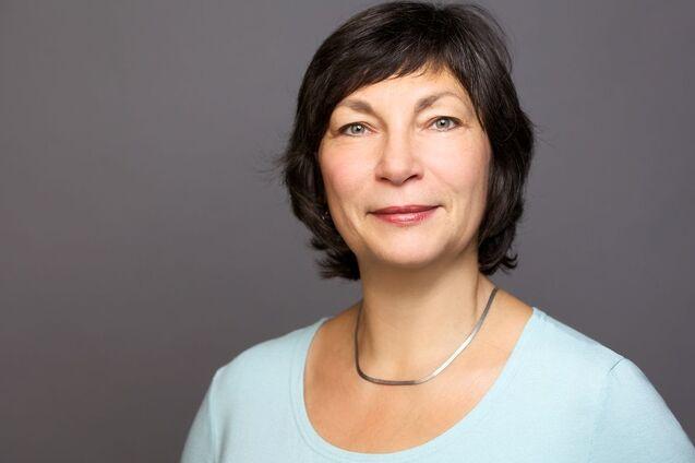 Porträt einer Frau mit kurzen, dunklen Haaren und einer blauen Bluse, auf grauem Hintergrund. Sie lächelt neutral.