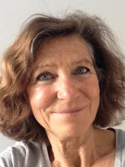 Portrait einer lächelnden Frau mit kurzem, lockigem Haar vor neutralem Hintergrund. Sie trägt ein graues Oberteil.