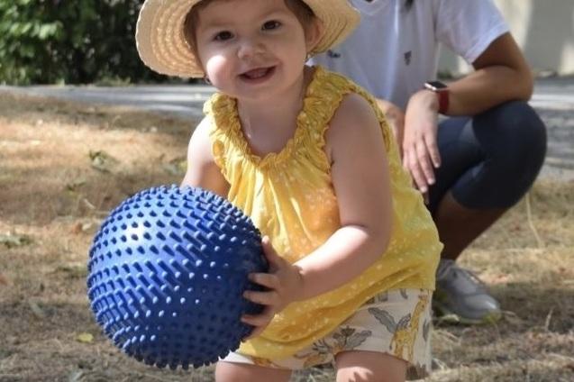 Lächelndes Kleinkind mit blauer Kinderball in der Hand, während eine Frau im Hintergrund zusieht.