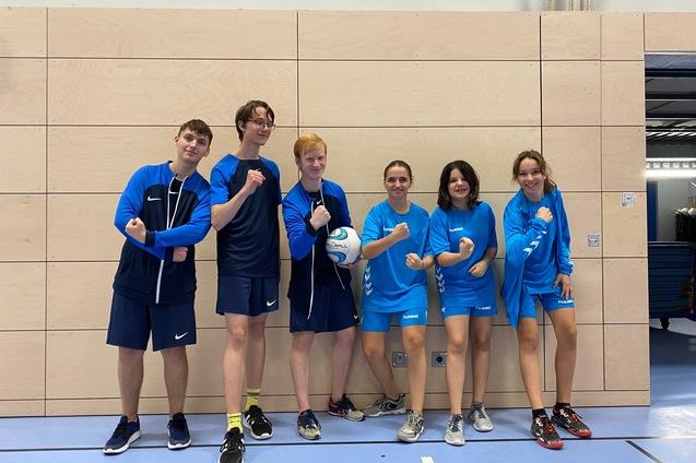 Sechs Jugendliche in blauen Sportoutfits posieren gemeinsam lächelnd mit einem Fußball vor einer Holzwand.