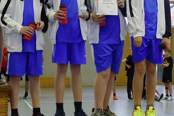 Vier Jugendliche in blauen Sportanzügen stehen auf einem Podium und halten Pokal und Urkunde. Hintergrund einer Sporthalle.