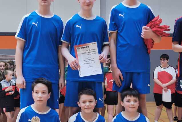 Sechs Jungen in blauen Sporttrikots stehen und sitzen in einer Sporthalle, der eine hält einen Pokal und der andere ein Zertifikat.