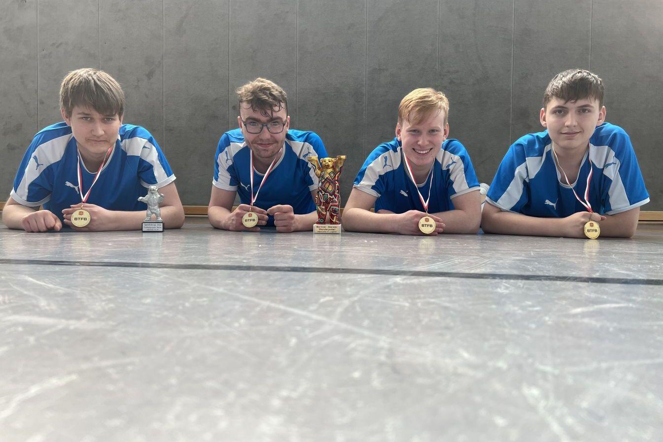 Vier Jugendliche in blauen Sporttrikots liegen auf einem Tischtennistisch mit Medaillen und Pokalen vor ihnen.