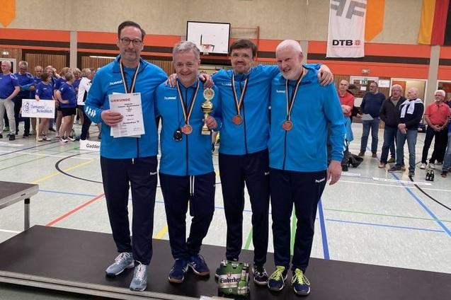 Vier Männer in blauen Sportjacken stehen auf einem Podium, halten Medaillen und einen Pokal, während Zuschauer im Hintergrund applaudieren.